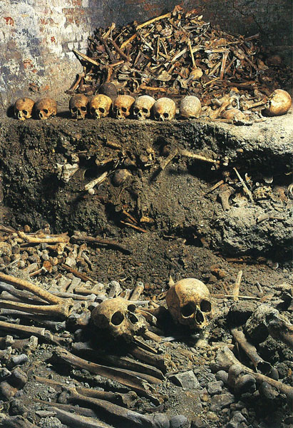 Charnel House showing many skulls and a large pile of bones. Other bones can be seen in the dirt.